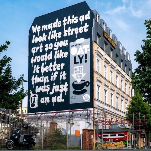 Black and white Oatly billboard in Berlin. The text is dripping as if just painted on, reading: "We made this ad look like street art so you would like it better than if it was just an ad." There is a paint can with paint brush sticking out of the top in the bottom left corner and there is a picture of their oat milk carton to the right of the text.